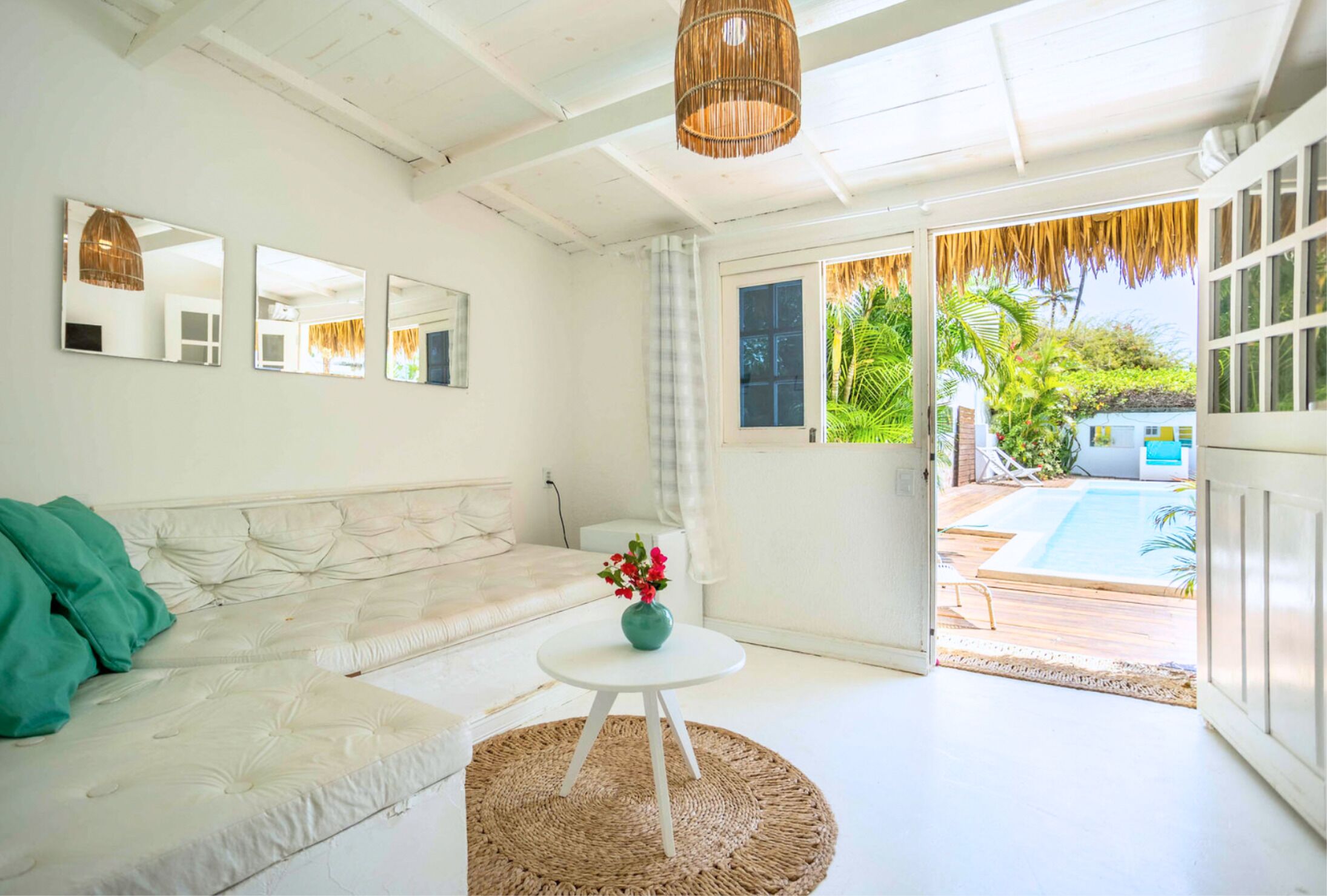 A bright living room with a white sofa, a round table with a vase, and a view of a pool outside.
