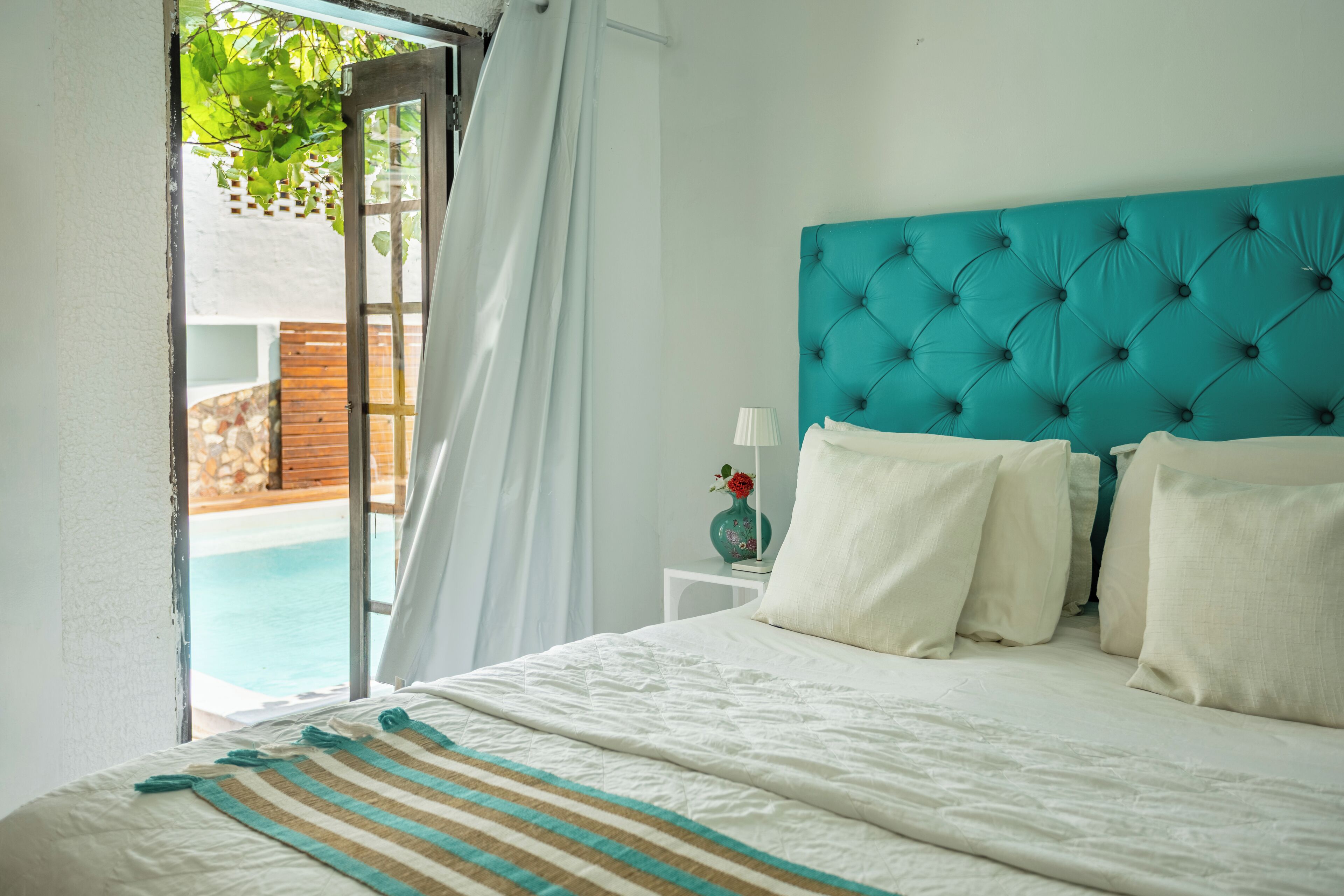 Une chambre avec un grand lit, une tête de lit de couleur turquoise, du linge de lit blanc, une petite table de chevet avec une lampe, et une vue sur une piscine par une fenêtre ouverte.