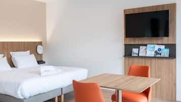 Una habitación de hotel moderna con una cama, una mesa de madera con dos sillas naranjas, un televisor de pantalla plana montado en la pared y un pequeño estante con folletos.