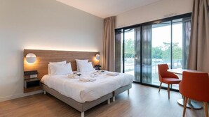 A modern hotel room with a large bed, a bedside table, a chair, and a view of the outdoors through a large window. - Villa Thermae Nancy (Nancy)