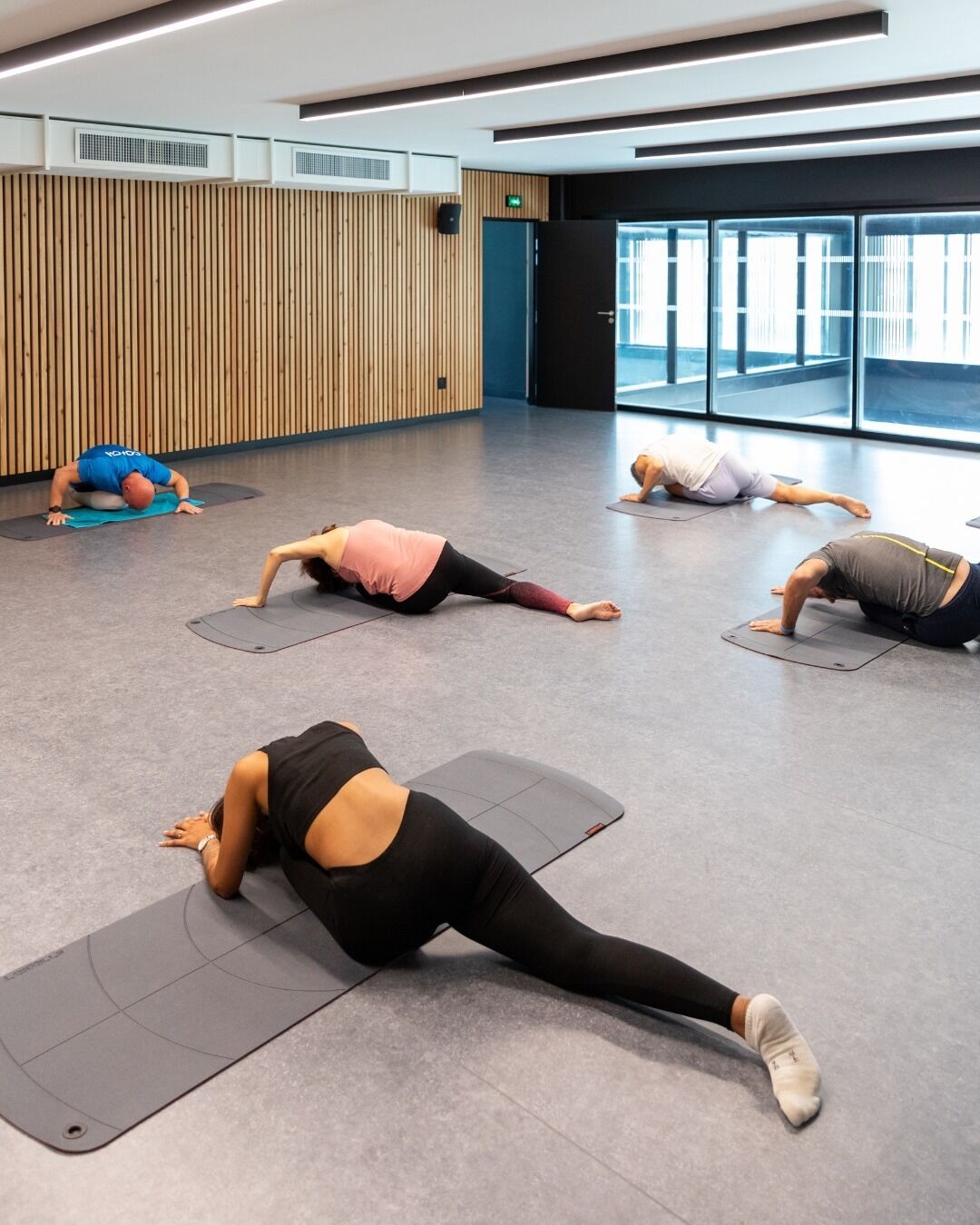 Ein geräumiges Zimmer mit großen Fenstern, einer Holzwand und mehreren Personen, die auf Matten Yoga praktizieren.