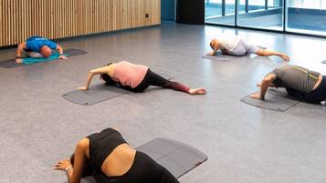Ein geräumiges Zimmer mit großen Fenstern, einer Holzwand und mehreren Personen, die auf Matten Yoga praktizieren.