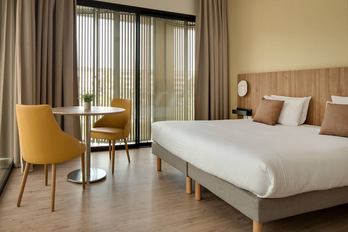 Una habitación de hotel con una cama grande, una mesa redonda con dos sillas y vistas a través de ventanales de gran altura.