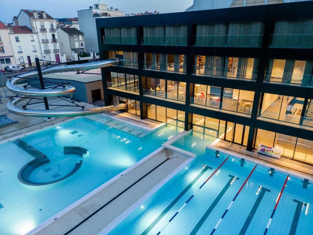An indoor swimming pool with multiple lanes, a water slide, and a modern building with large windows.