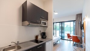 A modern kitchen with dark cabinets, a stainless steel sink, and a microwave. There is a small refrigerator, a coffee maker, and a window overlooking a balcony with orange chairs. - Villa Thermae Nancy (Nancy)