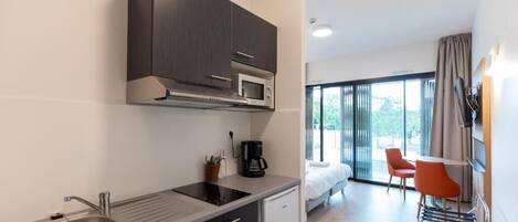 A modern kitchen with dark cabinets, a stainless steel sink, and a microwave. There is a small refrigerator, a coffee maker, and a window overlooking a balcony with orange chairs.