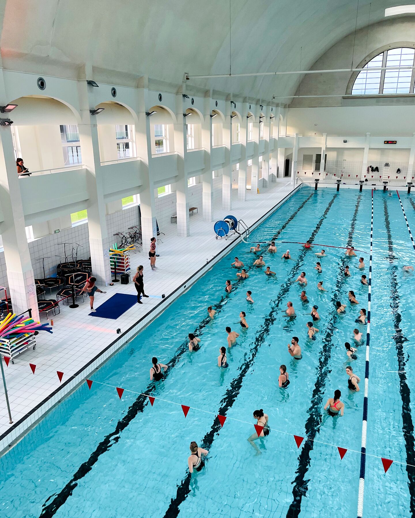 A large indoor swimming pool with multiple lanes, a spectator area with chairs, and a roofed upper level with windows.