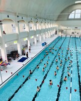 Una gran piscina cubierta con múltiples carriles, una zona para espectadores con sillas y un nivel superior cubierto con ventanas.