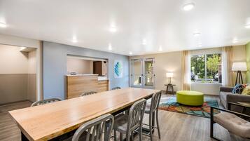 Une chambre d’hôtel avec une table à manger en bois, des chaises grises, un pouf vert, un canapé, une fenêtre avec des rideaux et un comptoir de réception.
