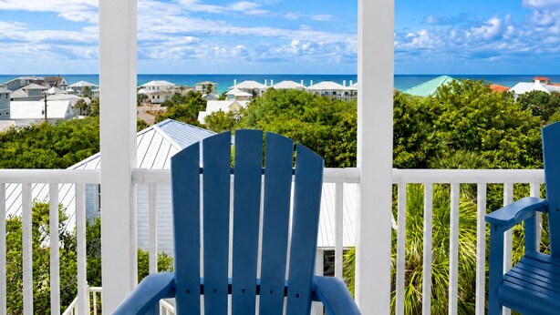 Ein Balkon mit zwei blauen Stühlen, weißen Geländern und Blick auf eine Küstenstadt mit Häusern und dem Meer.