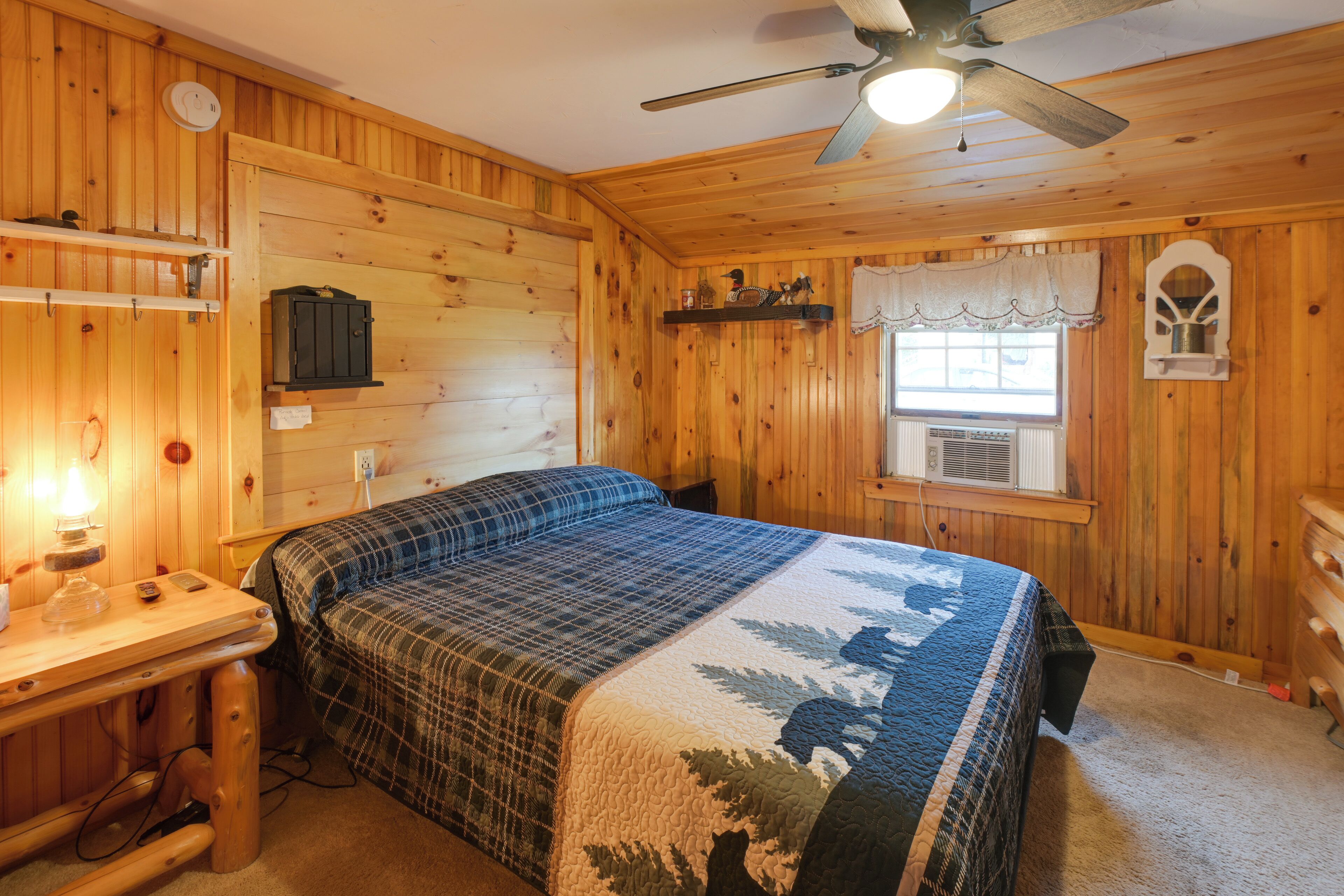 A wooden cabin room with a bed, a desk, a lamp, and a ceiling fan.
