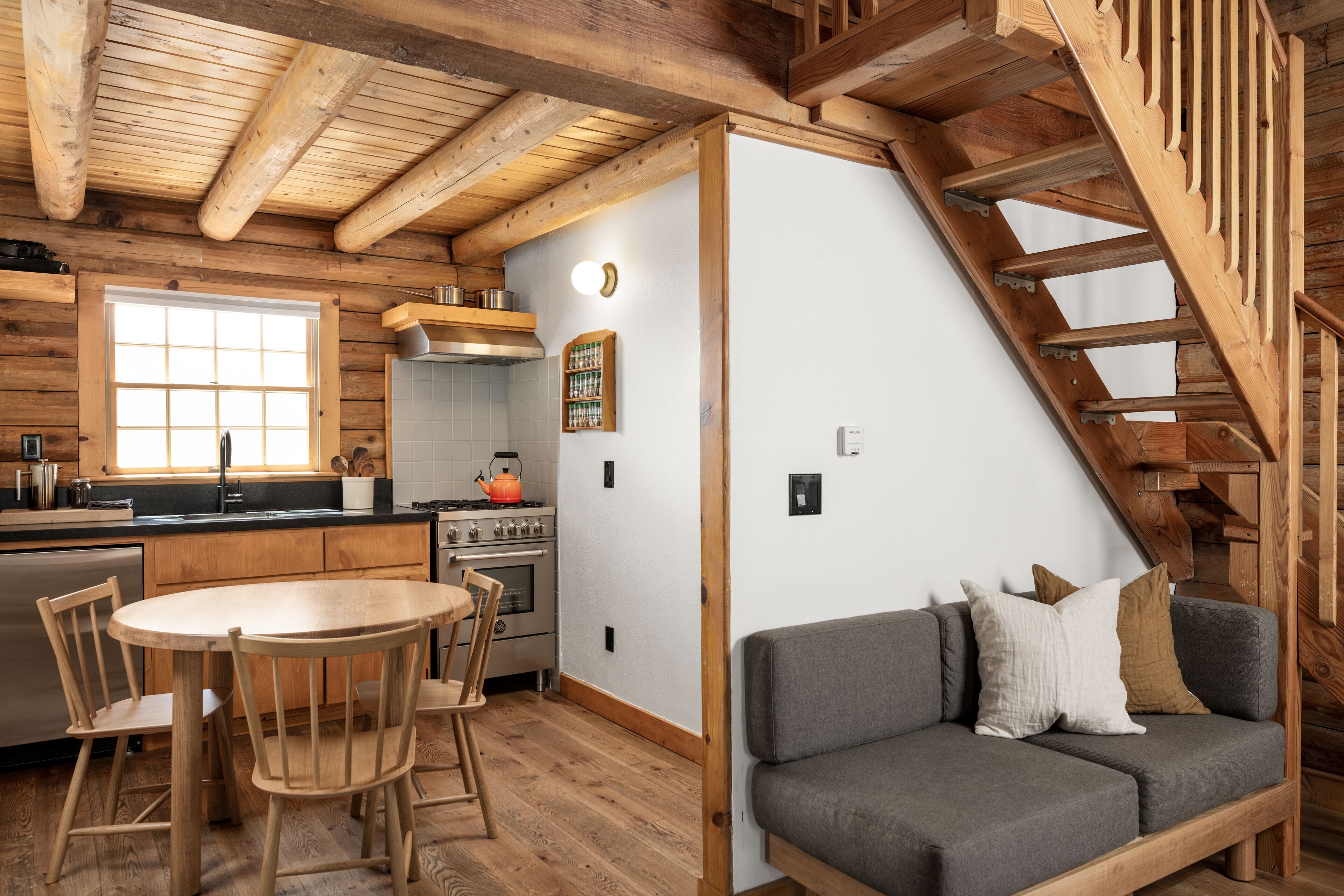 Un acogedor interior de cabaña con una mesa de comedor de madera, sillas, un sofá gris, una cocina con estufa y una escalera de madera.