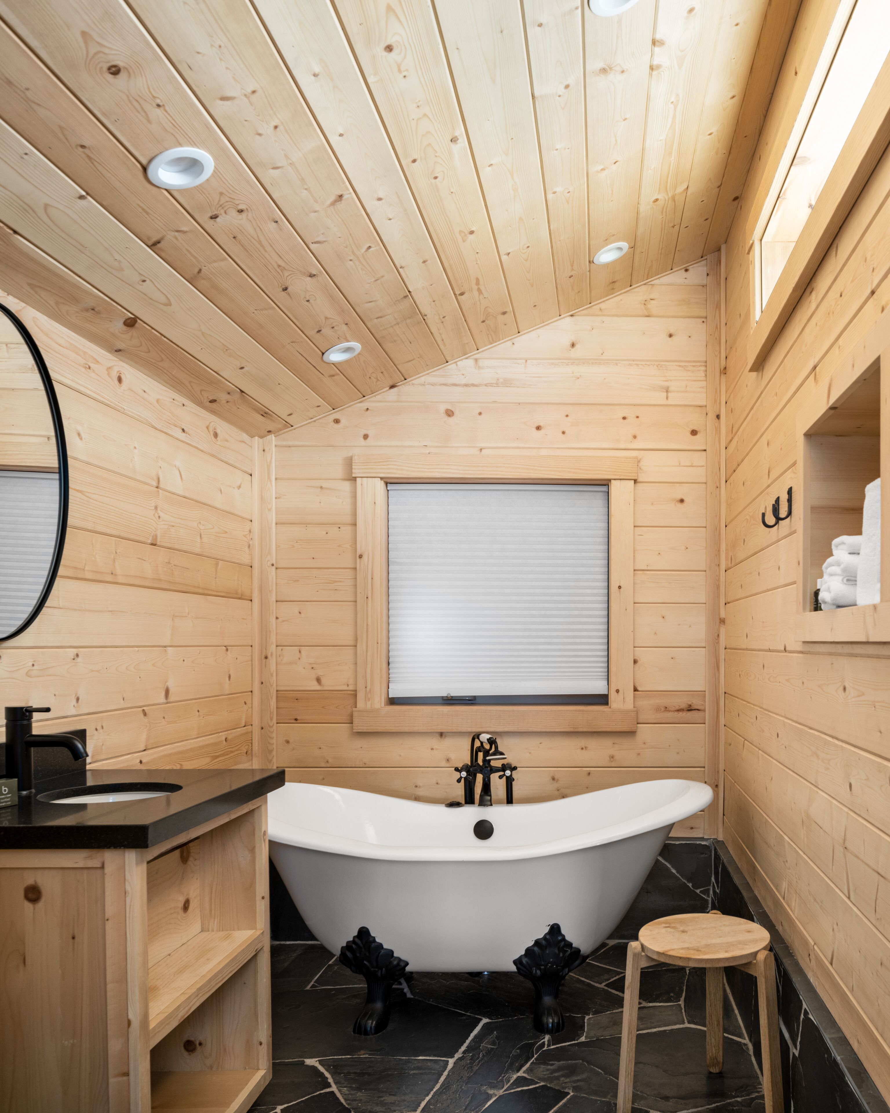 Une salle de bain rustique, avec des murs en bois, une baignoire sur pieds, un tabouret en bois et une fenêtre équipée de stores.