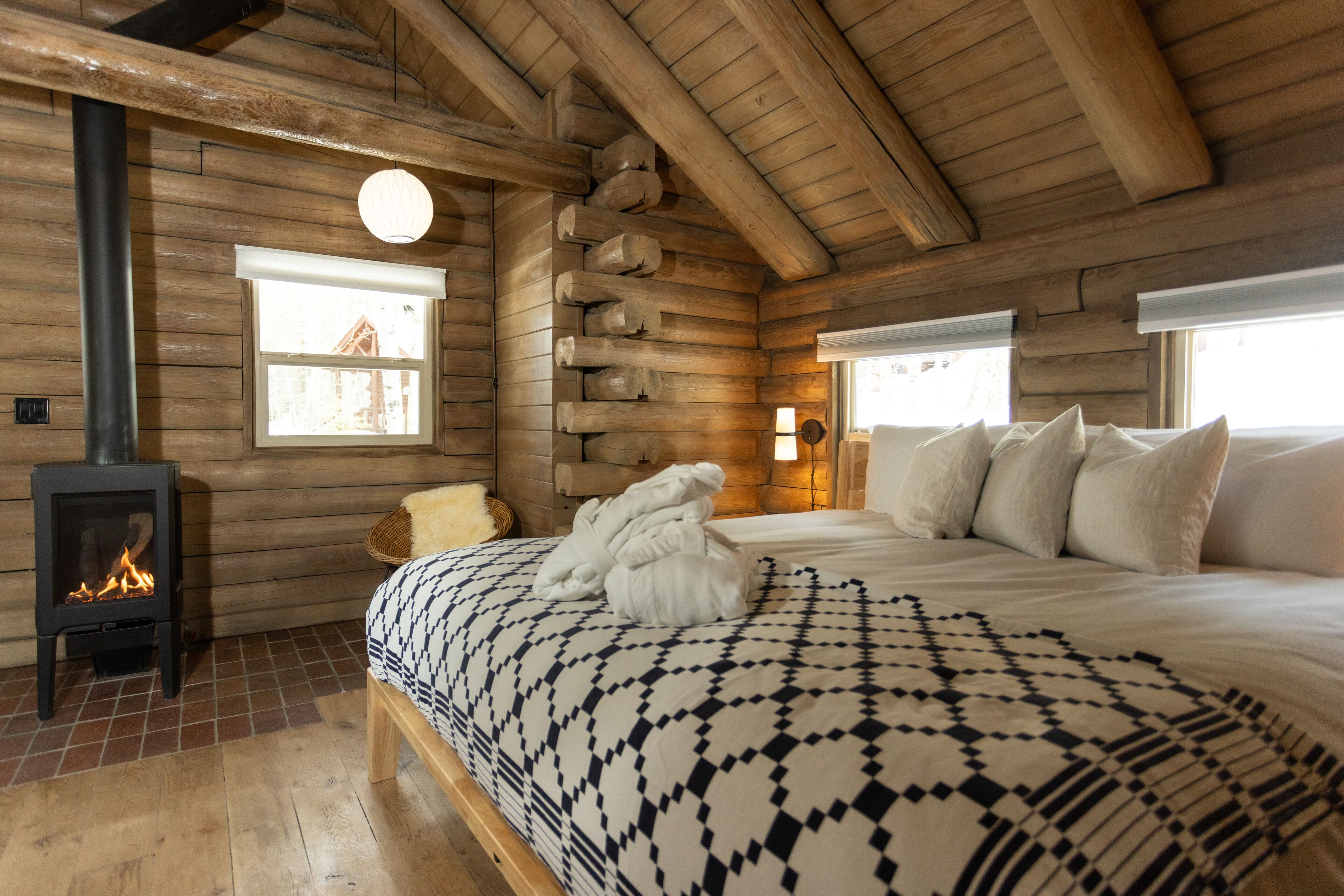 A log cabin bedroom with a fireplace, a bed with patterned bedding, and a large window.