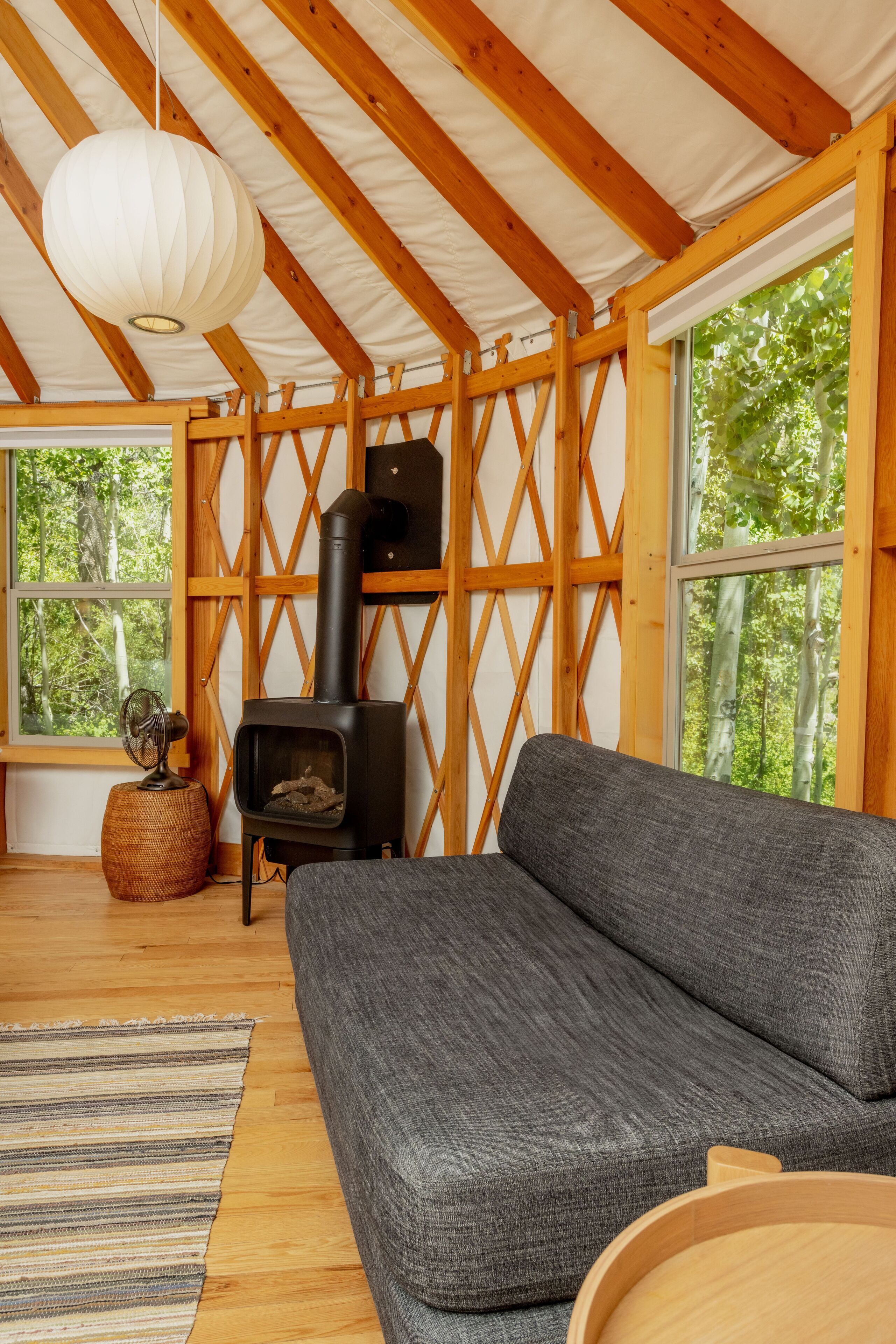 A cozy room with a gray sofa, a wooden ceiling, a black wood stove, and large windows offering a view of trees.