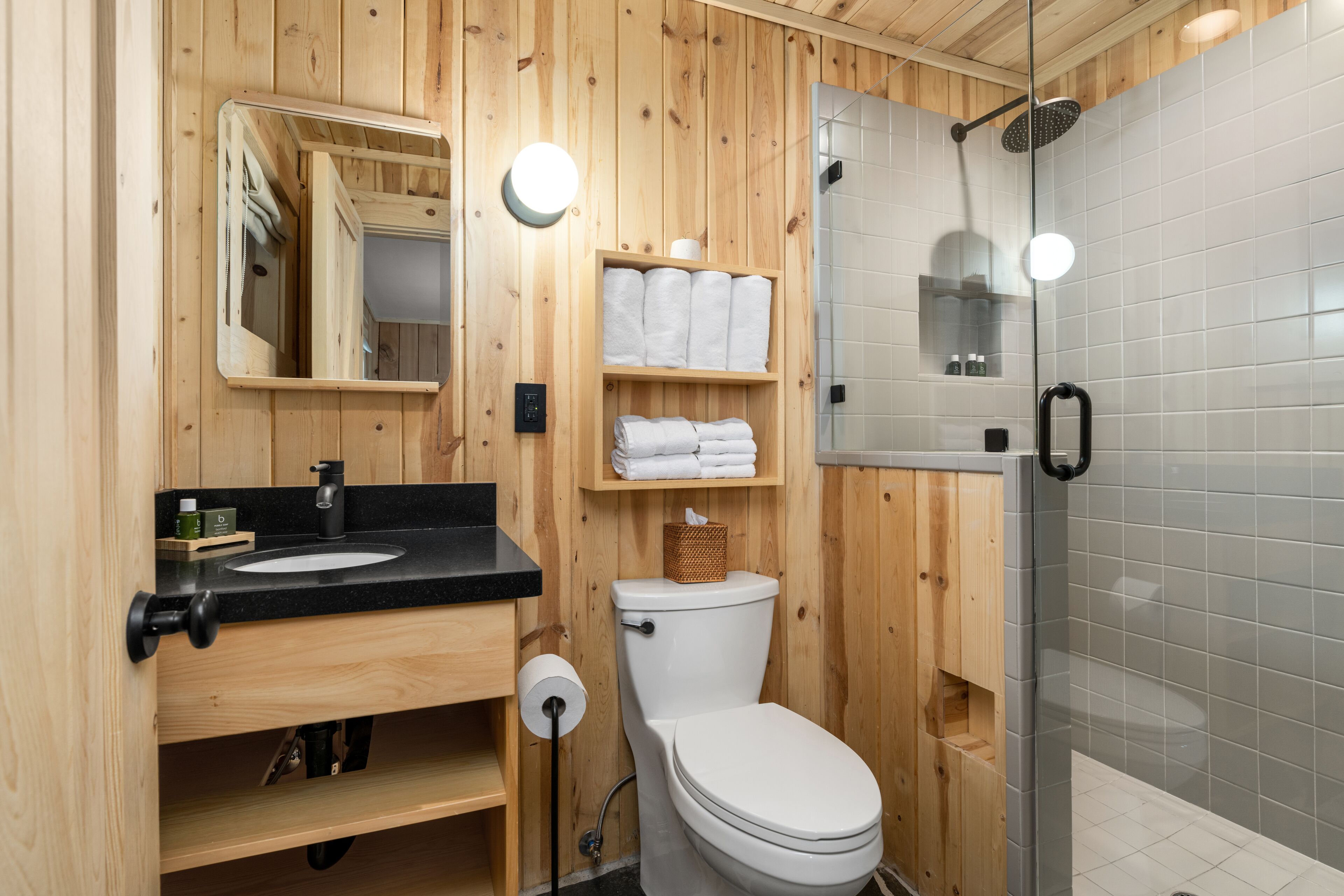 Un baño con paneles de madera, un lavabo negro, un inodoro blanco y una ducha con cerramiento de vidrio.