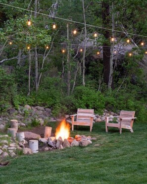 An outdoor seating area with two wooden chairs, a fire pit, and string lights.
