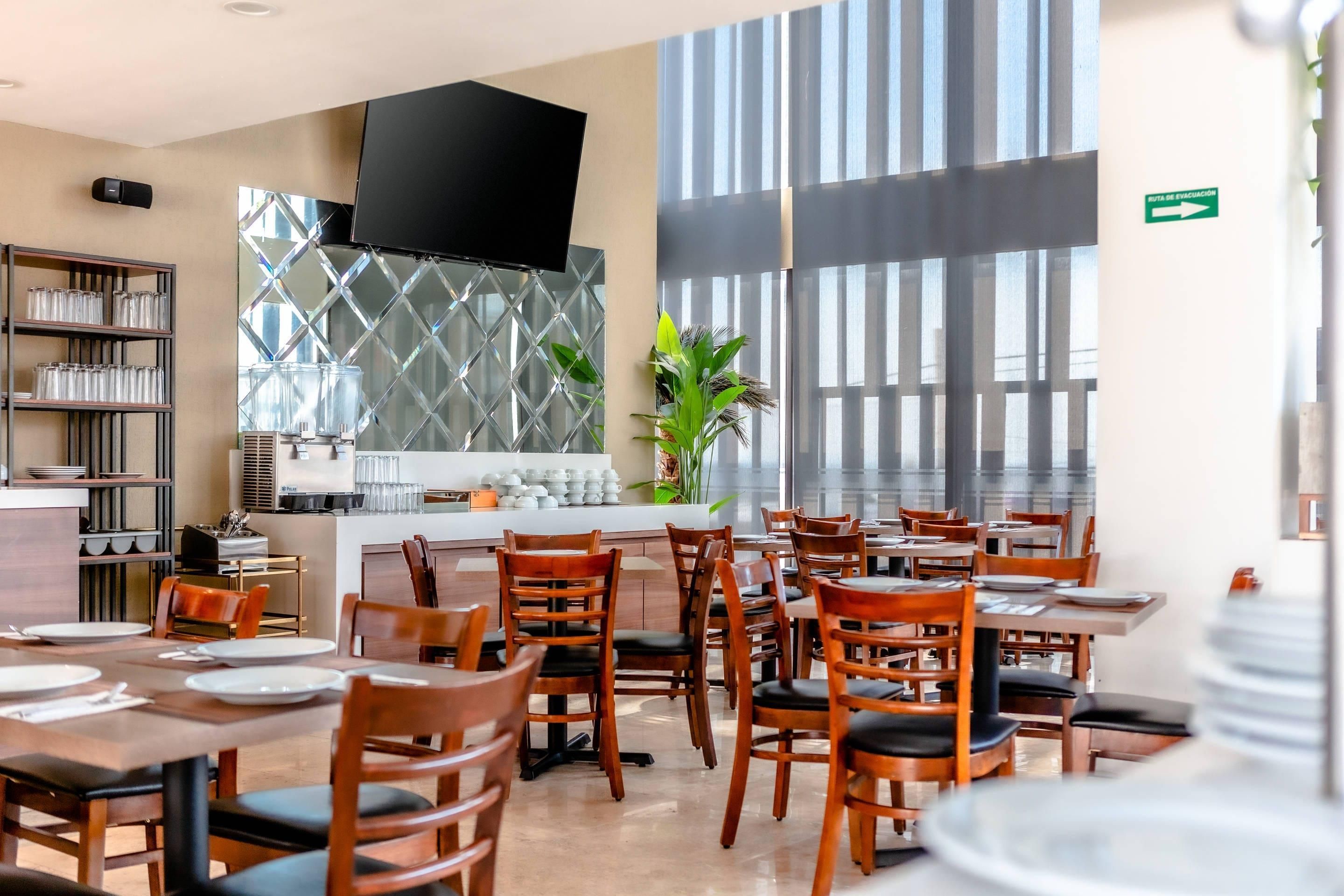 Un restaurante con mesas y sillas de madera, un televisor en la pared y un ventanal con persianas.