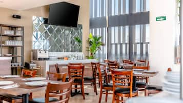 Un restaurante con mesas y sillas de madera, un televisor en la pared y un ventanal con persianas.