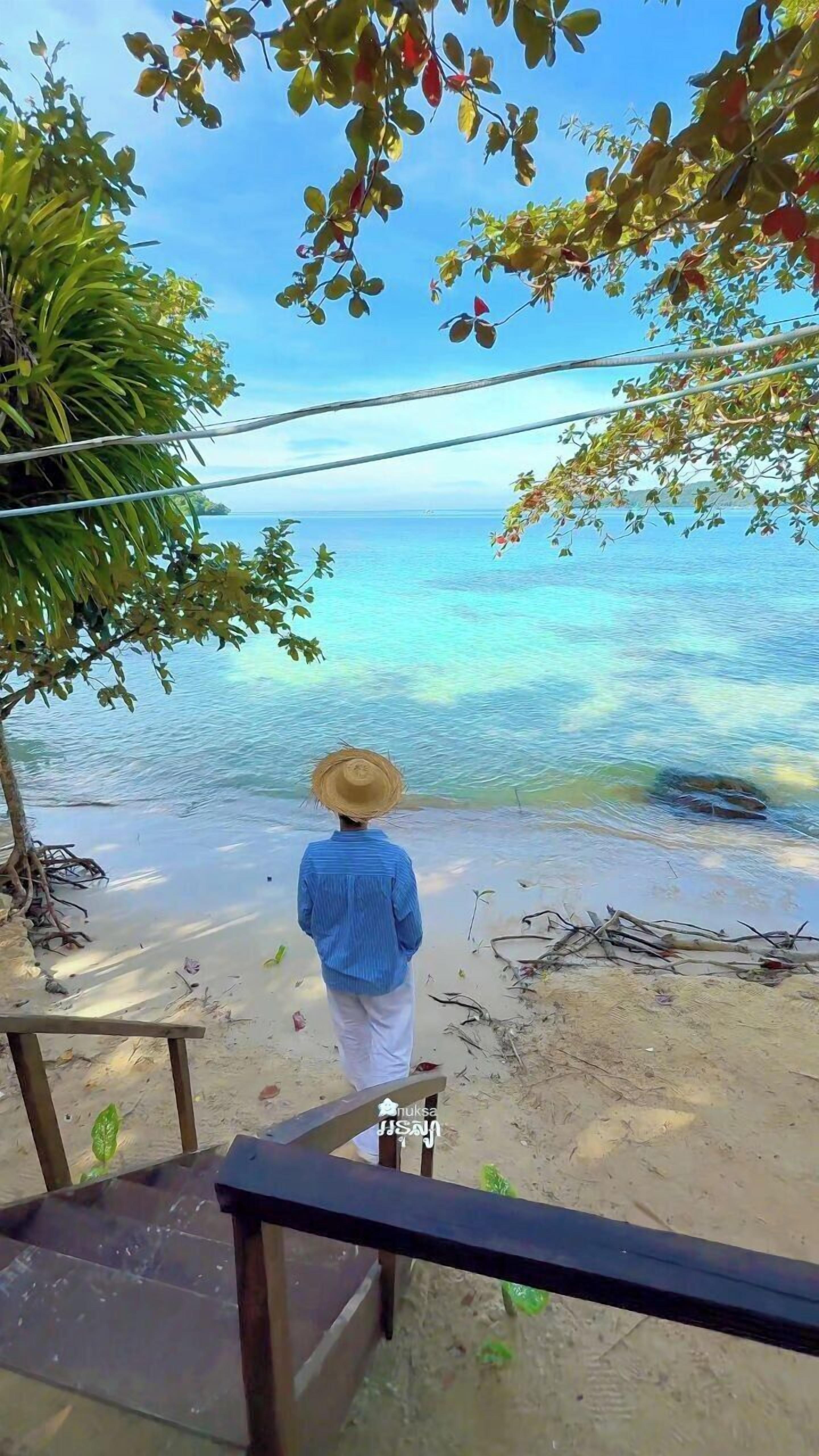 Una persona con un cappello di paglia e una camicia blu in piedi su una spiaggia sabbiosa con vista sull'oceano.