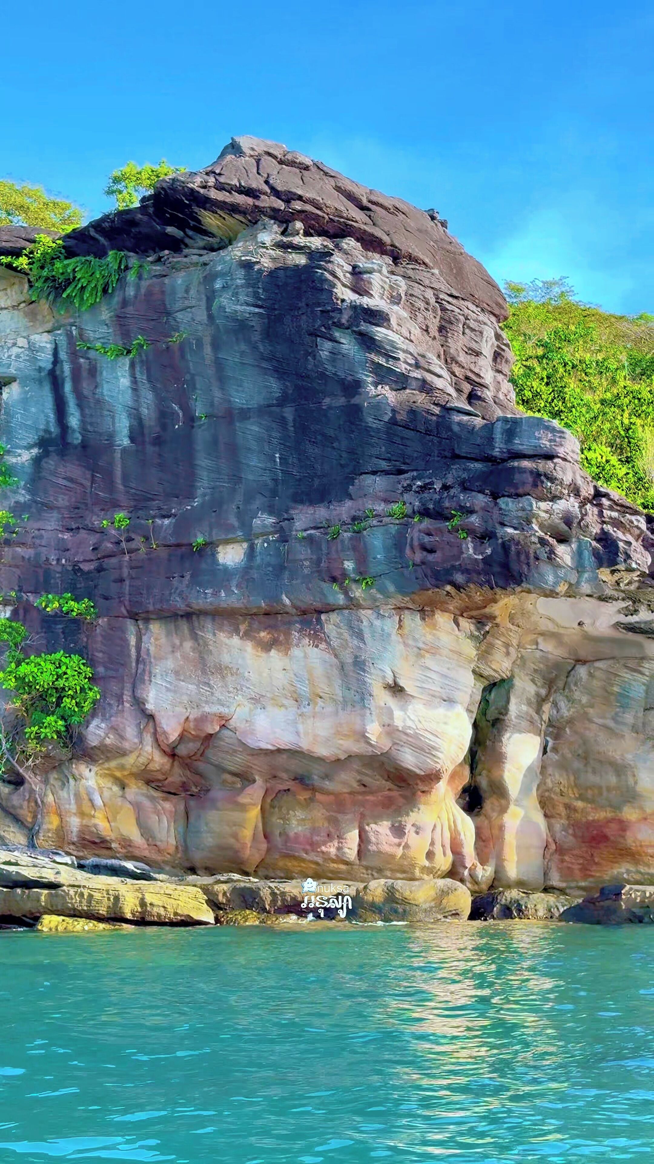 Imponente formazione rocciosa con l'ingresso di una grotta, circondata da acque cristalline e vegetazione.