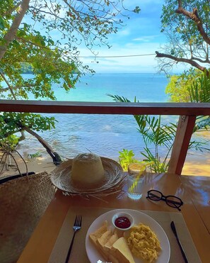 A table with a plate of scrambled eggs, toast, and ketchup, a straw, and a hat, with a view of the sea and trees in the background. - Koh Ta Kiev Island Resort (Koh Ta Kiev Island)