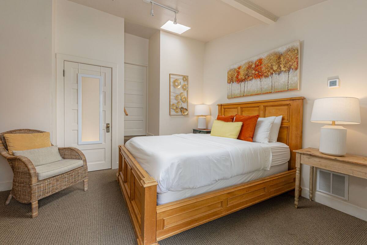 A bedroom with a wooden bed, a wicker chair, a bedside table with a lamp, and a painting on the wall.