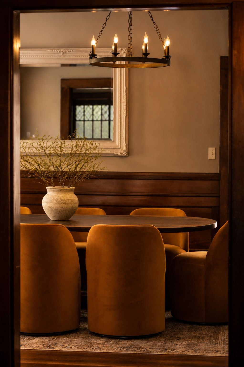 A dining area with a round table and velvet chairs, a chandelier, and a vase with plants.