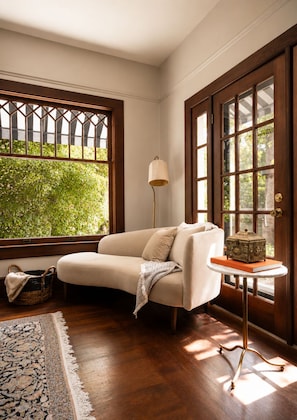 A room with a beige sofa, a floor lamp, a small table with a decorative box, and a large window with a view of greenery. - The De La Vina Inn (Santa Barbara)