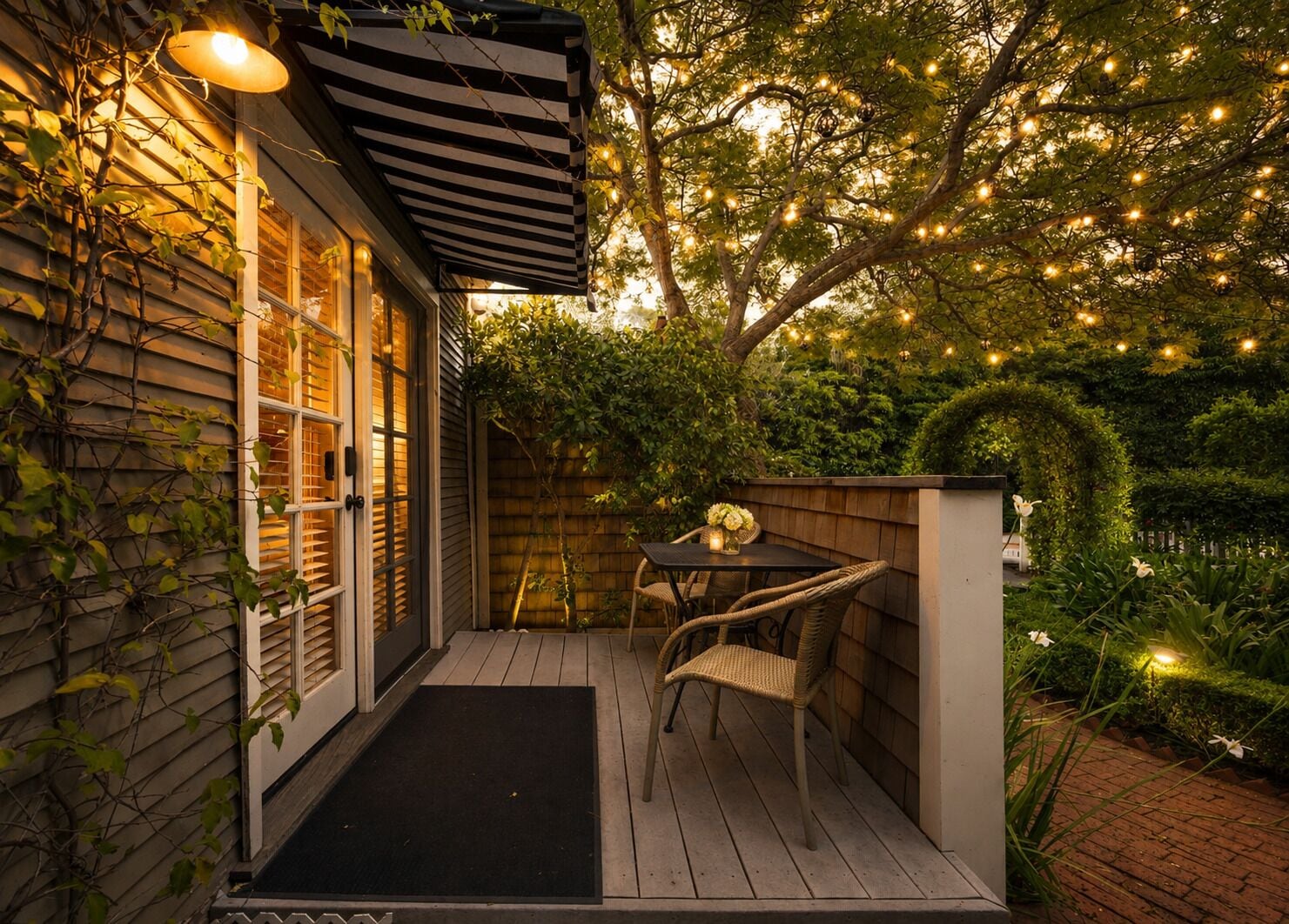 Une terrasse en bois avec une table et des chaises, entourée de verdure et illuminée de guirlandes lumineuses.