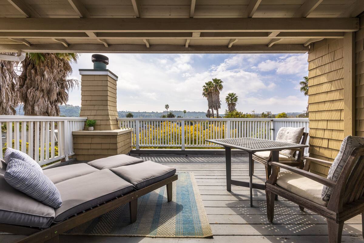 A covered outdoor seating area with a view of the ocean, a white fence, and palm trees.