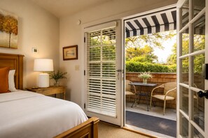 A hotel room with a bed, a nightstand with a lamp, a framed picture, and a sliding glass door leading to an outdoor seating area with chairs and a table. - The De La Vina Inn (Santa Barbara)