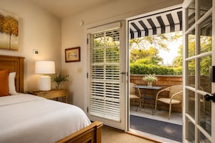 Habitación de hotel con cama, mesita de noche con lámpara, un cuadro enmarcado y puerta corrediza de vidrio que da a un área exterior con sillas y una mesa.