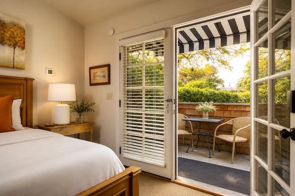 A hotel room with a bed, a nightstand with a lamp, a framed picture, and a sliding glass door leading to an outdoor seating area with chairs and a table. - The De La Vina Inn (Santa Barbara)