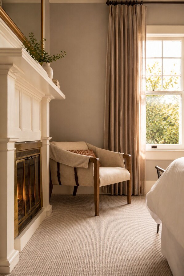 A room with a fireplace, a chair, a window with curtains, and a view of greenery outside. - The De La Vina Inn (Santa Barbara)