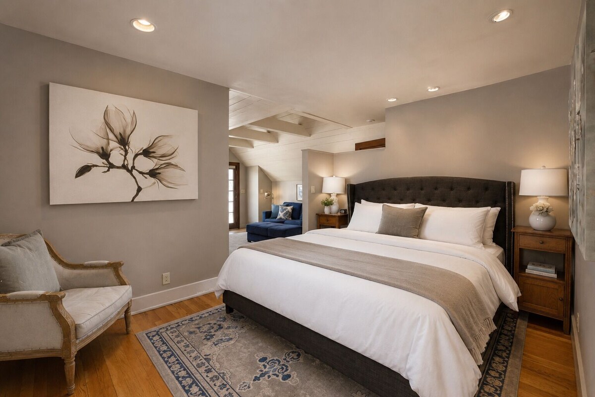 Quarto com cama grande, uma cadeira, mesa de cabeceira com abajur e um quadro pendurado na parede.