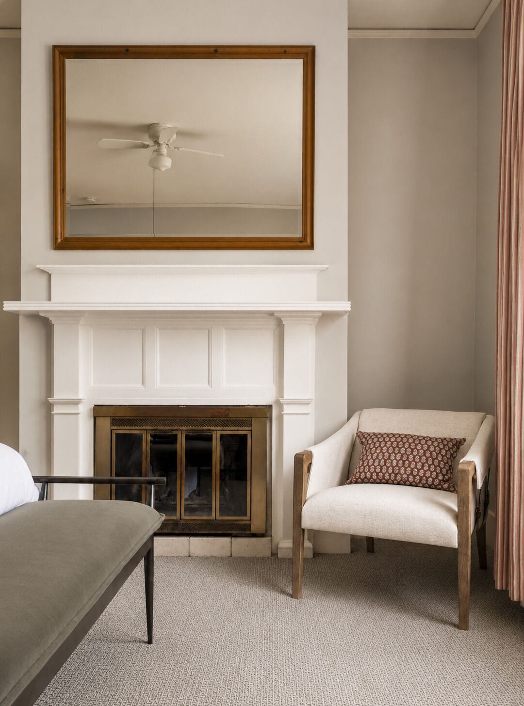 A room with a white fireplace, a ceiling fan, a chair with a patterned pillow, and a bed with a grey headboard.