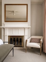 Una habitación con chimenea blanca, ventilador de techo, silla con cojín estampado y cama con cabecera gris.