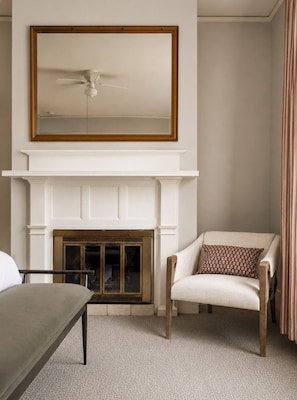 A room with a white fireplace, a ceiling fan, a chair with a patterned pillow, and a bed with a grey headboard. - The De La Vina Inn (Santa Barbara)