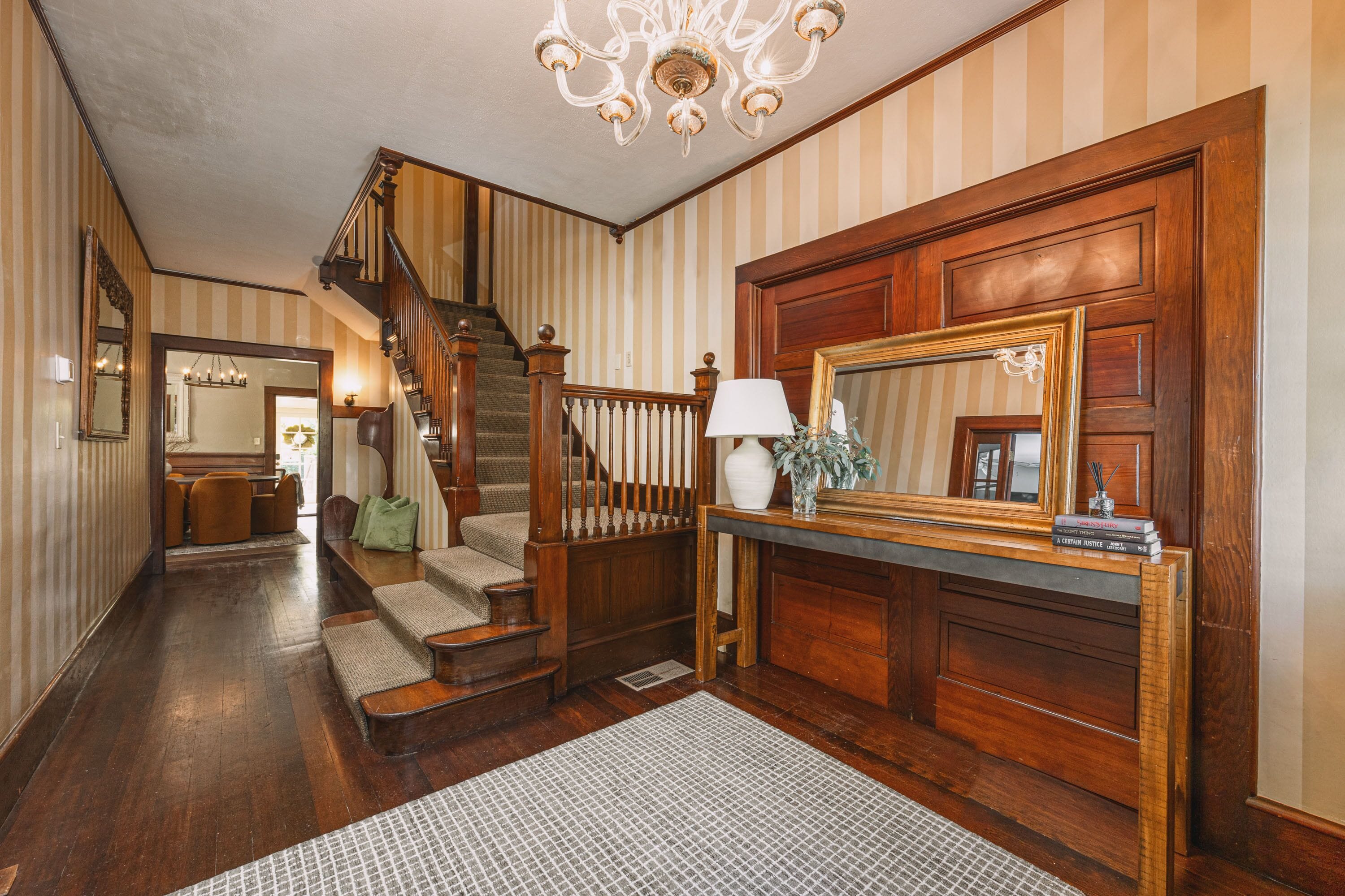 A hallway with a wooden staircase, a mirror, a lamp, and a fireplace.