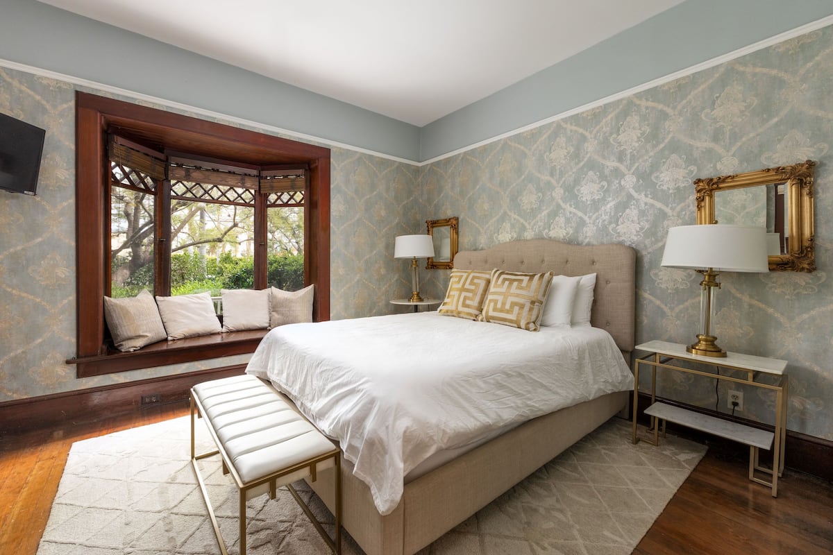A bedroom with a large bed, a bench, a nightstand, and a window with a view of greenery.