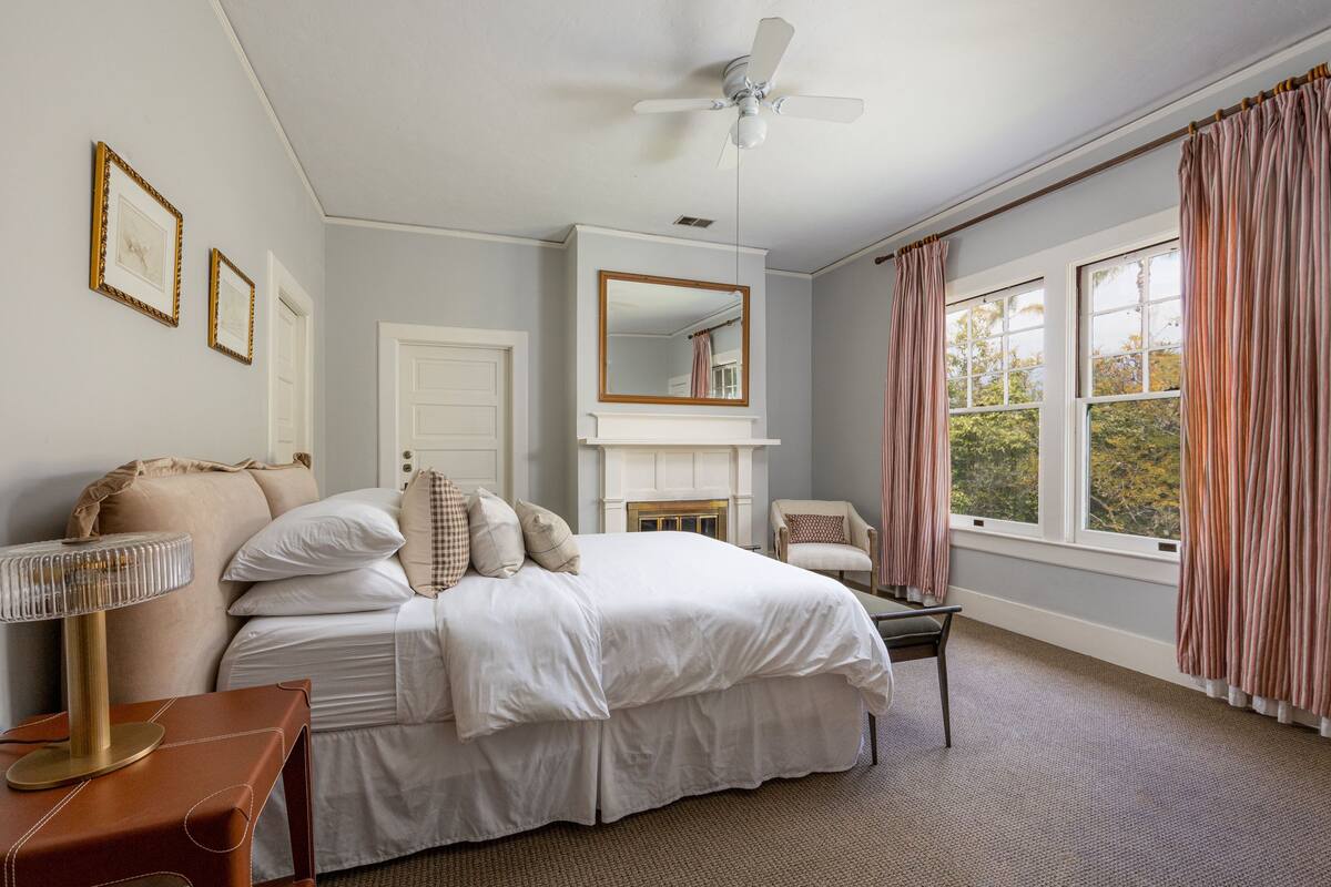 A bedroom with a bed, a chair, a mirror, a fireplace, and a window with curtains.