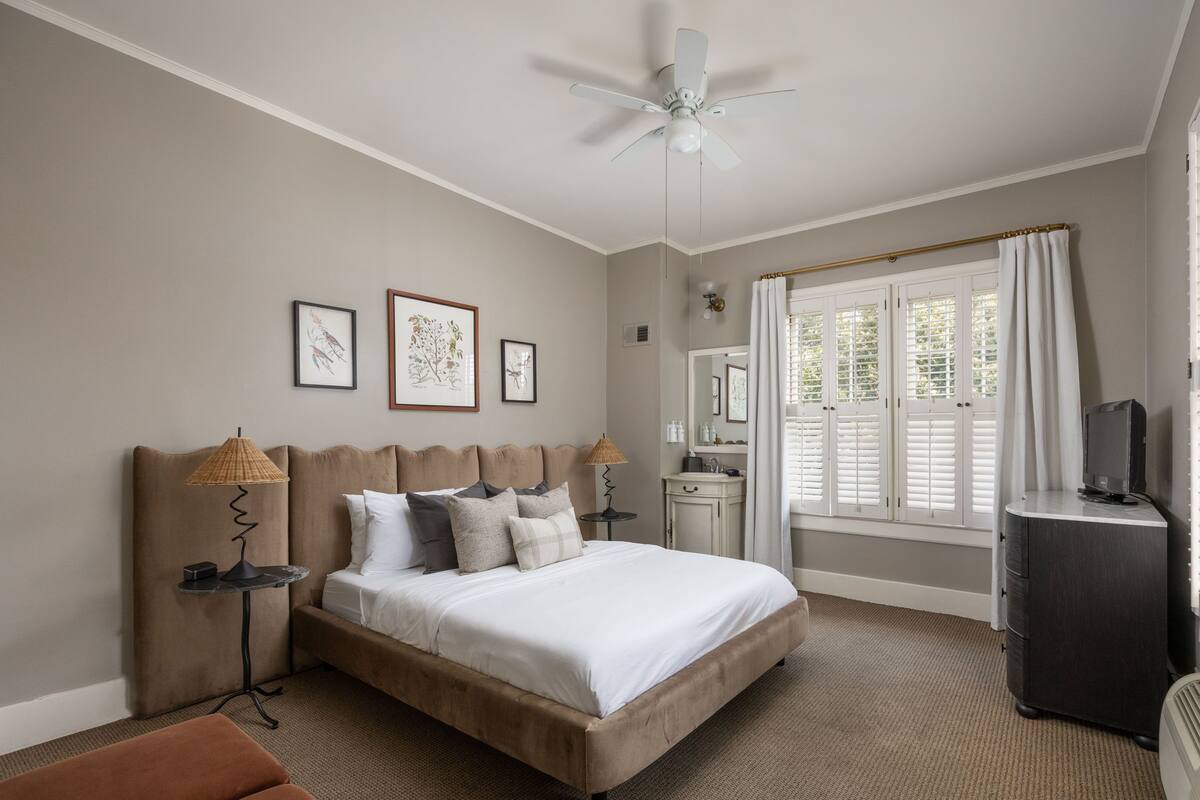 A bedroom with a bed, bedside lamps, a ceiling fan, a dresser, a TV, and a window with shutters.