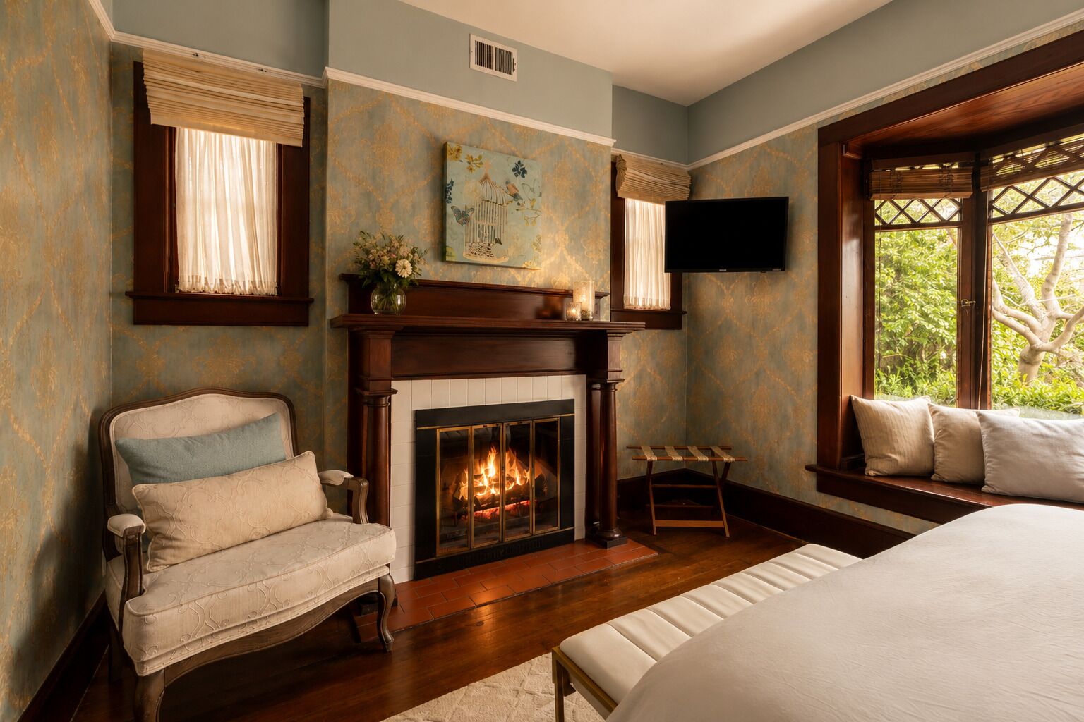 A bedroom with a fireplace, a bed, a chair, and a window with a view of greenery.