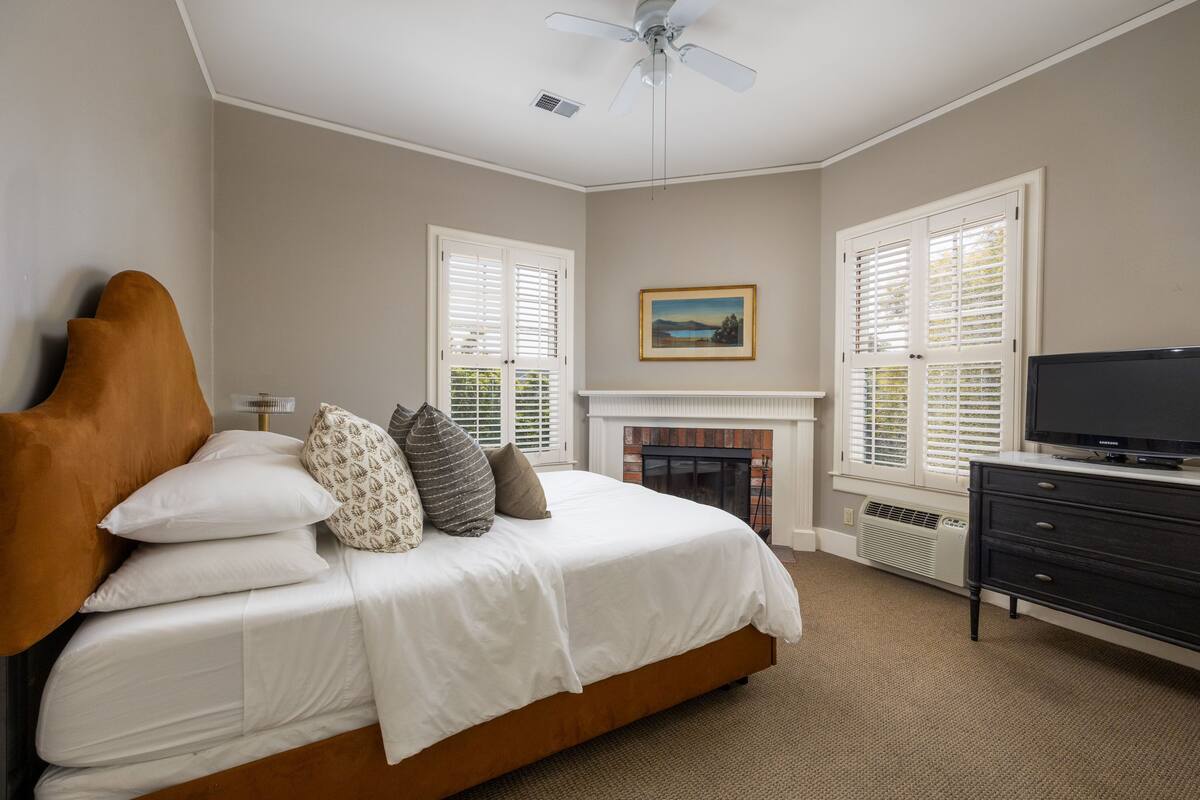 A bedroom with a bed, a television, a fireplace, and windows with shutters.
