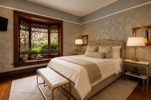 A bedroom with a large bed, two bedside tables, a bench, and a window with a view of greenery.