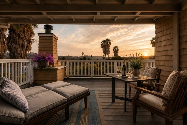 A covered outdoor patio with a wooden table and chairs, a cushioned bench, and a view of palm trees and a sunset. - The De La Vina Inn (Santa Barbara)
