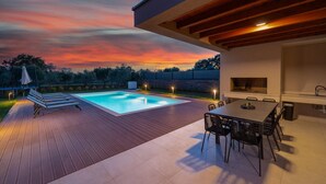 Ein Schwimmbad mit Holzterrasse, Grillplatz und Blick auf einen Sonnenuntergang.