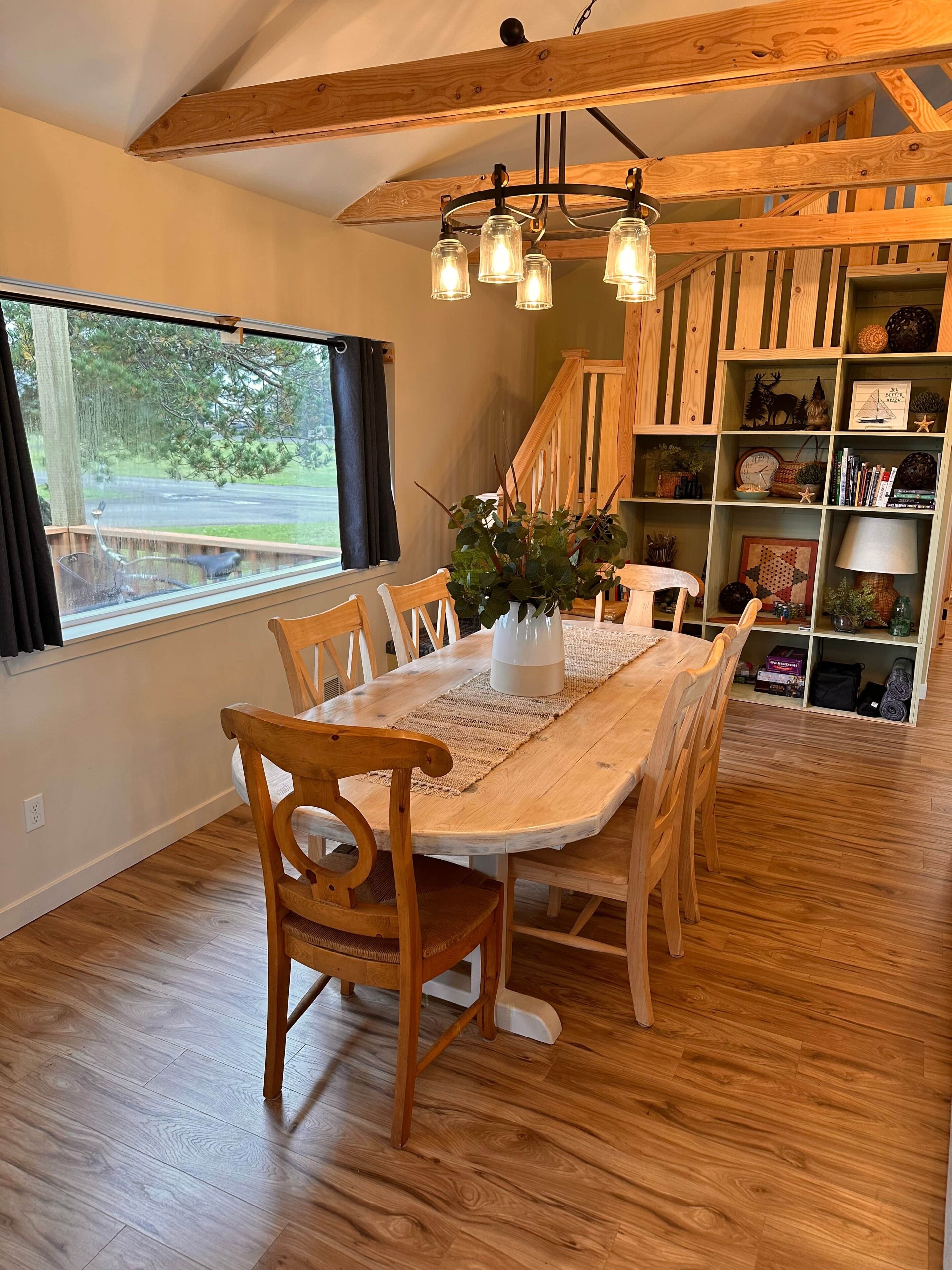 A dining area with a wooden table and chairs, a chandelier, and a bookshelf with various items.