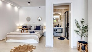 A modern bedroom with a bed, bedside tables, and a nightstand. There is a bathroom visible through an open door.
